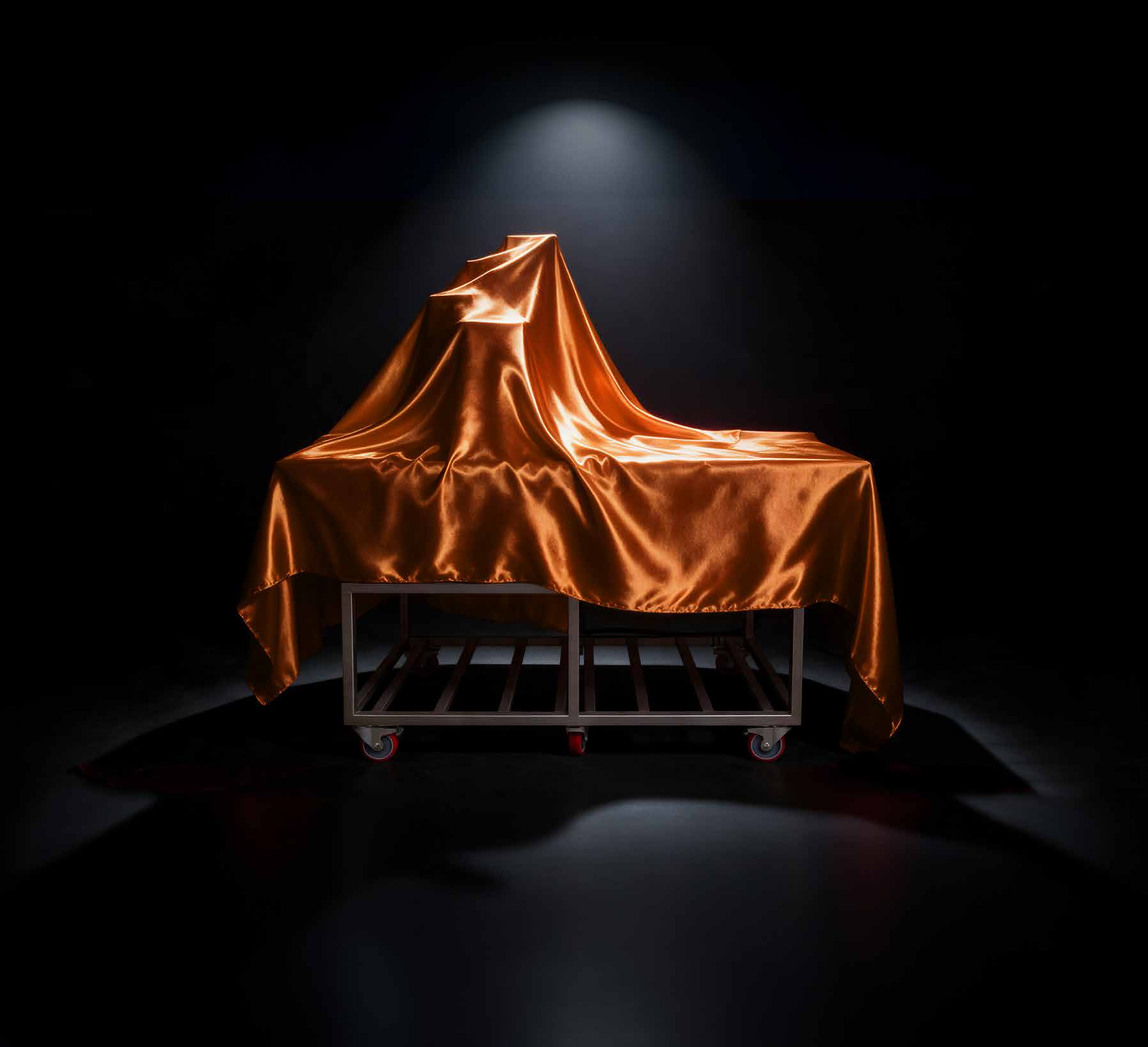 Een machine onder een glanzend oranje doek, uitgelicht door een spot in een donkere ruimte.